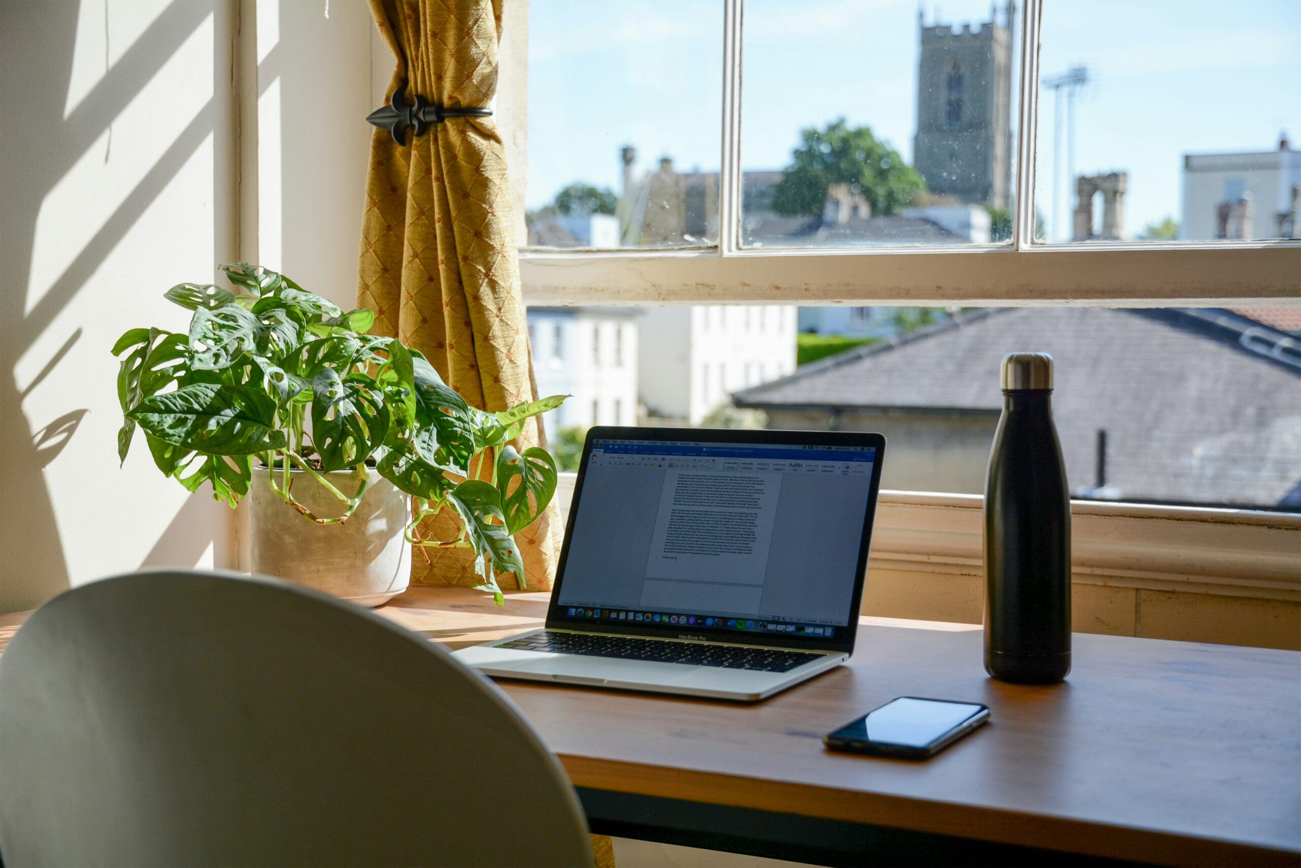 Laptop on a desk by a sunny window with a plant and notebook, representing freedom and flexibility for health practitioners building high-ticket programs online.
