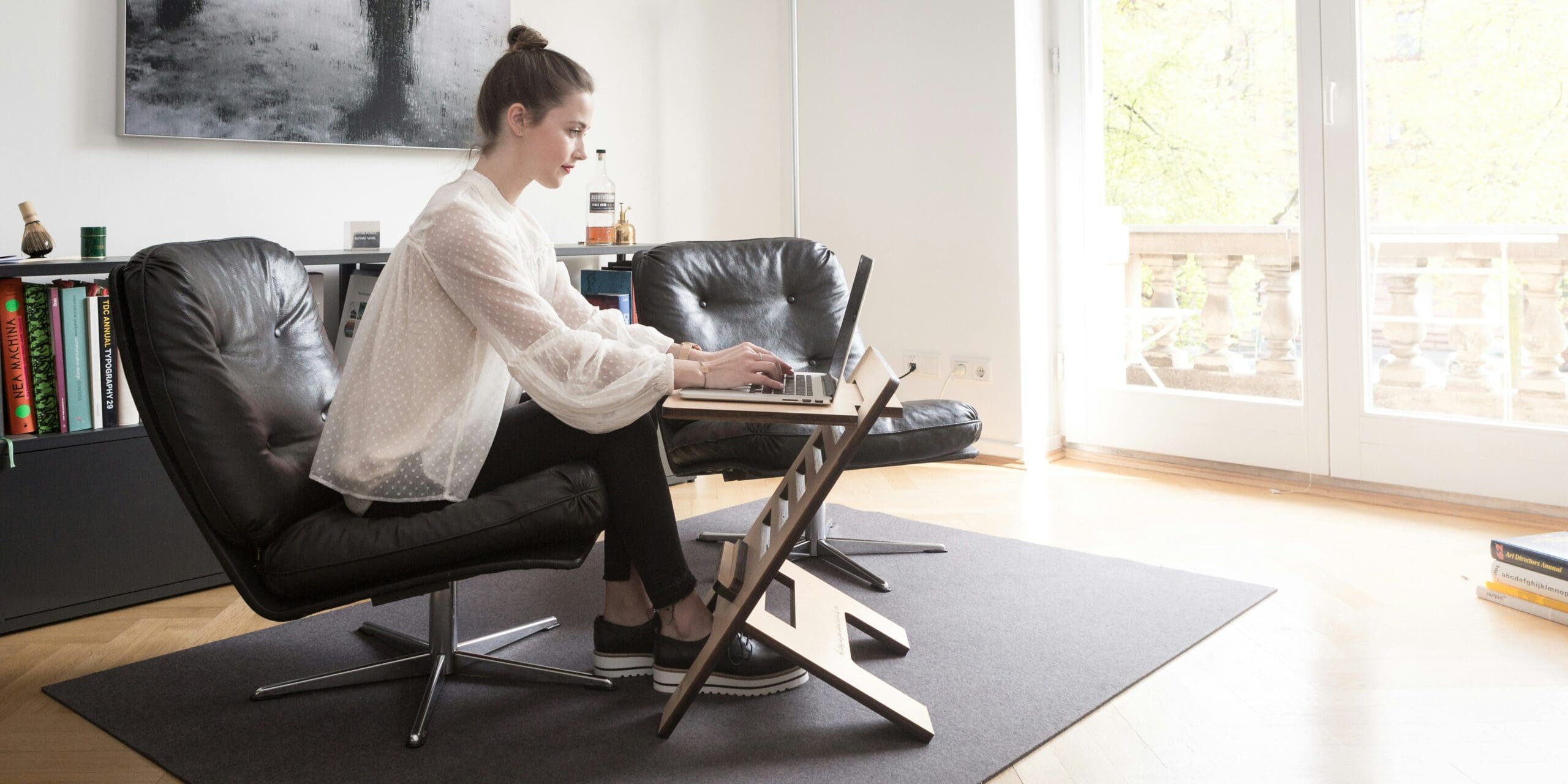 Woman working remotely on a laptop at home with a standing desk setup and bright natural light.